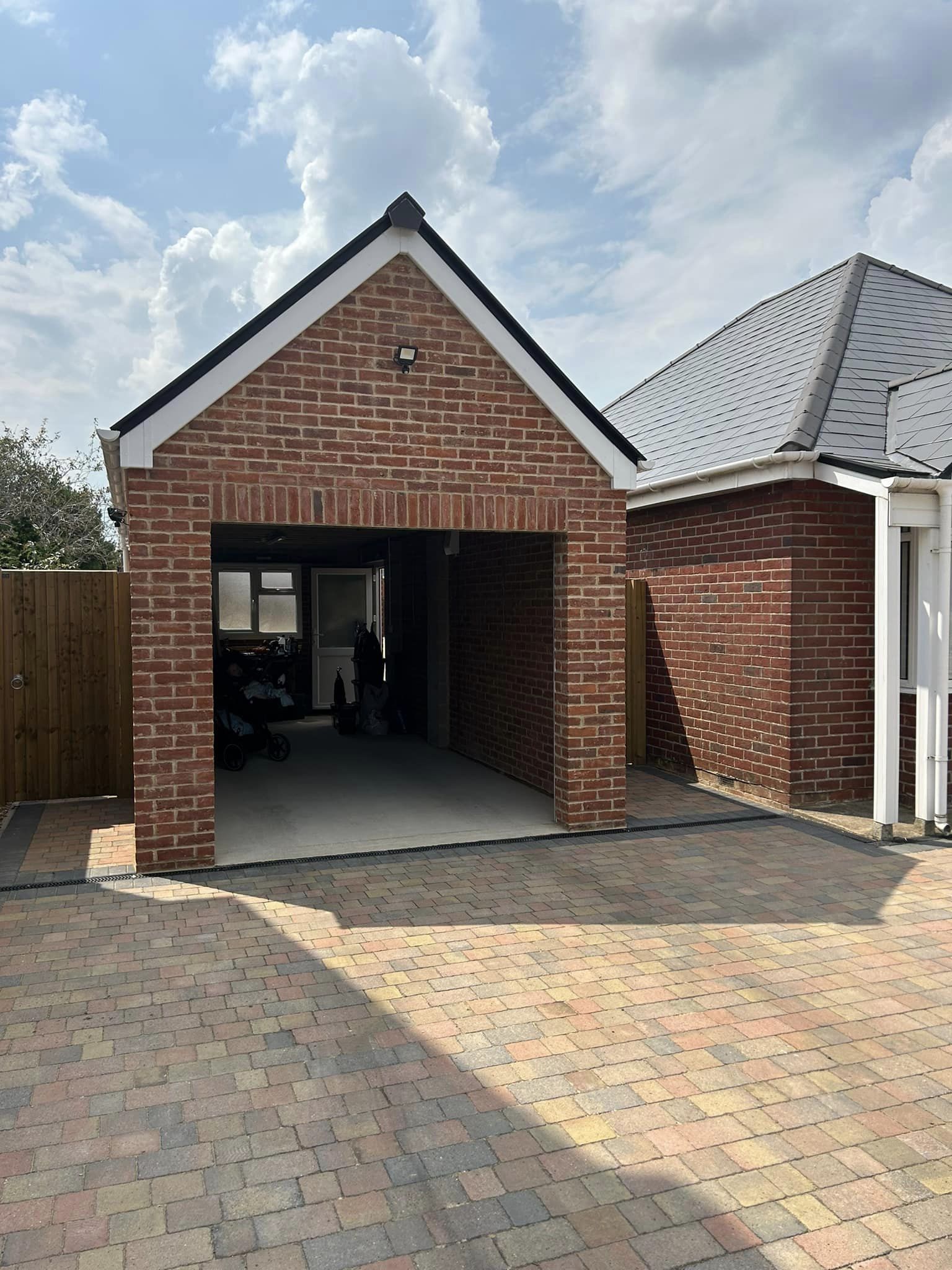 A brick garage with an open entrance is attached to a house. The driveway is paved with multicolored bricks. The sky is partly cloudy, and there is a wooden fence to the left of the garage.