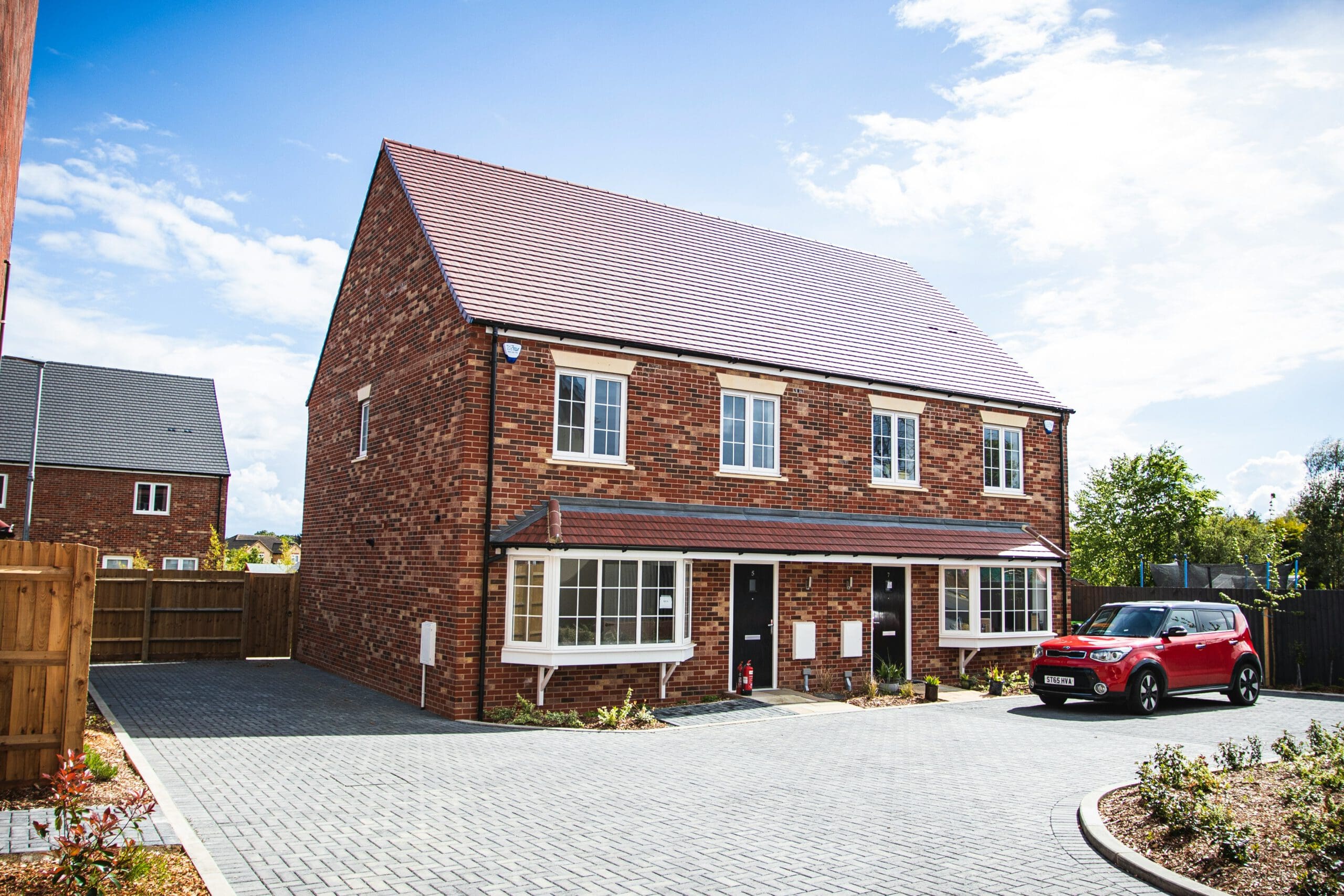 A modern, brick semi-detached house with a pitched roof and two bay windows. A red car is parked on the driveway, and the sky is clear and blue.