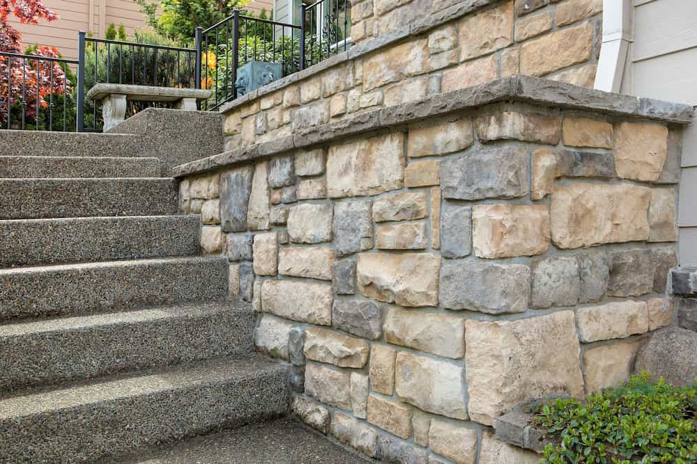 Exterior close-up of a house featuring textured stone veneer siding and a set of concrete stairs with a metal railing, surrounded by greenery and landscaping.